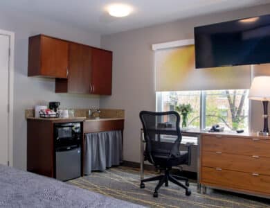 A hotel room corner with a desk, office chair, dresser, lamp, wall-mounted TV, and kitchenette area with a mini fridge, microwave, sink, and coffee maker.