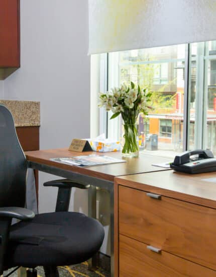 Modern office space with a black mesh chair, wooden desk, flower vase, telephone, documents, and a window overlooking a city street.