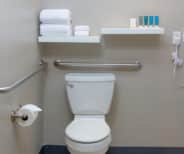 Accessible bathroom with a toilet, grab bars, toilet paper holder, stacked towels on shelves, and toiletries on a shelf above the tank.