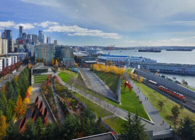 Aerial view of a city waterfront park with green lawns, walking paths, sculptures, nearby buildings, and a harbor with container cranes and water in the background.