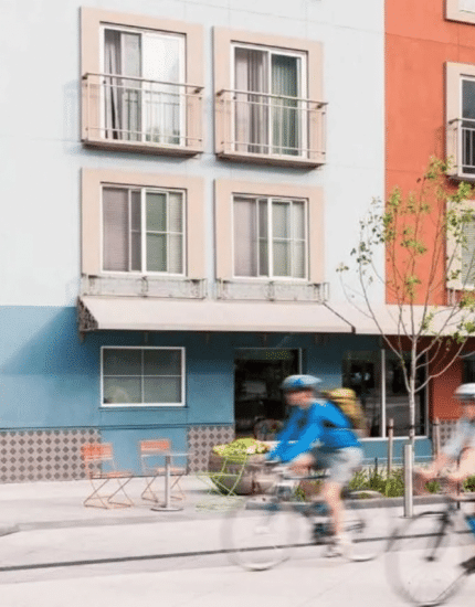 Four people ride bicycles past a colorful apartment building—one of the vibrant places to stay in Seattle, Washington—with a bus and trees visible in the background.