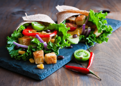 Two vegetable wraps with lettuce, tomato, cucumber, tofu, black beans, corn, and red onion on slate, garnished with a red chili pepper—a fresh option to enjoy while exploring Things To Do In Seattle.