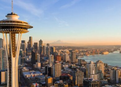 Aerial view of downtown Seattle with the Space Needle in the foreground, city buildings, waterfront, and Mount Rainier visible in the background under a clear sky.