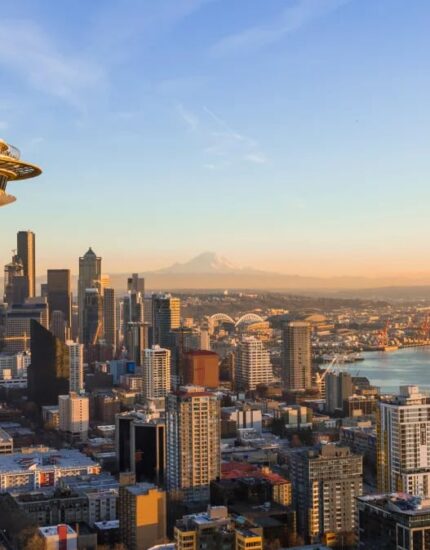 Aerial view of downtown Seattle with the Space Needle in the foreground, city buildings, waterfront, and Mount Rainier visible in the background under a clear sky.