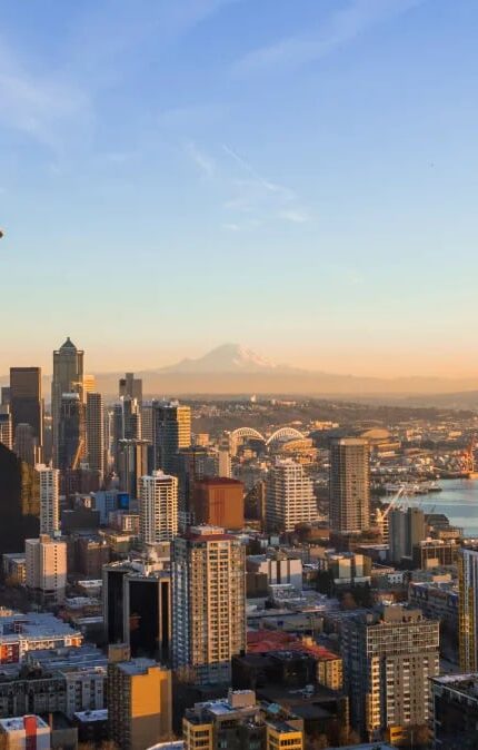 Aerial view of downtown Seattle with the Space Needle in the foreground, city buildings, waterfront, and Mount Rainier visible in the background under a clear sky.