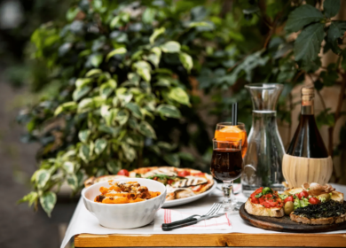 A wooden table set outdoors with dishes of pasta, grilled vegetables, bruschetta, and drinks creates the perfect ambiance—reminiscent of al fresco dining experiences among lush plants. It’s like discovering hidden gems while exploring Things To Do In Seattle.