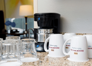 A coffee maker sits on a granite countertop next to several upside-down glass cups and white mugs labeled "Econo Lodge.