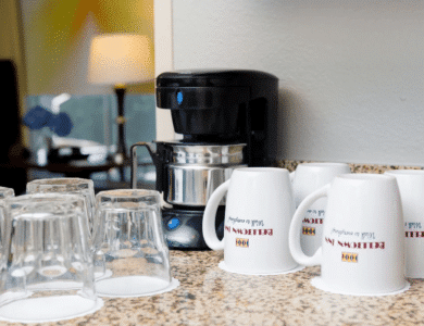 A coffee maker sits on a granite countertop next to several upside-down glass cups and white mugs labeled "Econo Lodge.
