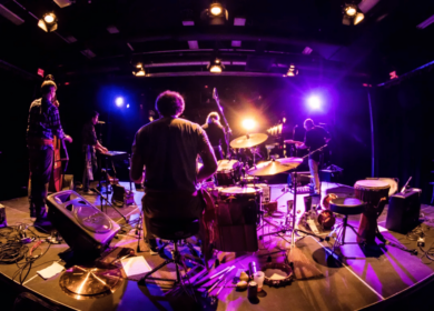 A band performs on stage under purple and yellow lights, with musicians playing drums, keyboard, and guitars, viewed from behind.