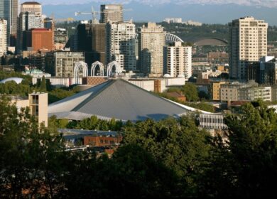 Downtown Seattle skyline with the distinctive sloped roof of Climate Pledge Arena in the foreground, surrounded by trees and high-rise buildings.