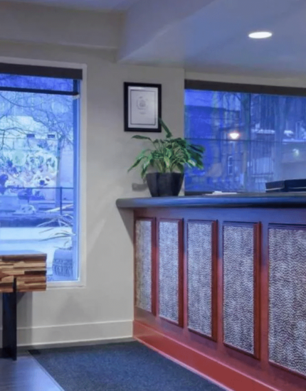 A hotel reception area with a red front desk, a small table with a flower vase, large windows, a plant, and wall art in the background.