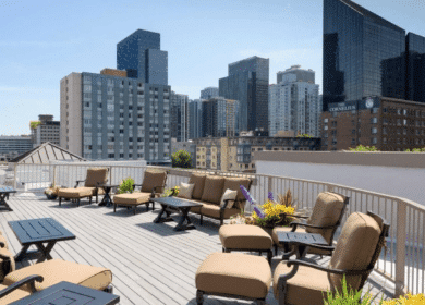 Rooftop patio with cushioned chairs and tables, potted plants, and a city skyline with modern high-rise buildings in the background.