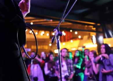 A musician performs on stage with a guitar; a microphone stand is visible, and a crowd of people watches in a dimly lit venue.