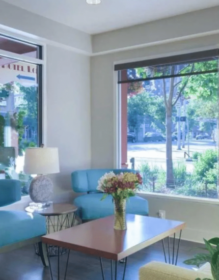 Modern waiting room with large windows, blue and yellow seating, a wooden coffee table with flowers, and ample natural light.