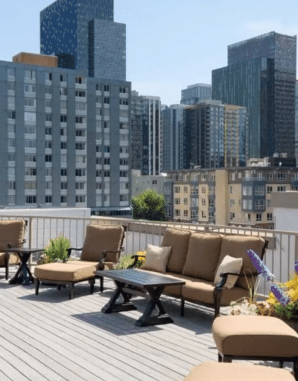 Outdoor rooftop patio with cushioned lounge chairs, tables, potted plants, and views of city high-rise buildings under a clear sky.
