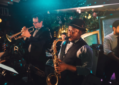 A jazz band performs on stage; a saxophonist is in the foreground, with a trumpeter and other musicians playing behind him under dim, colorful lighting.