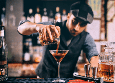 A bartender wearing a cap and vest pours a dark cocktail into a martini glass at a bar, surrounded by bottles and bar tools.