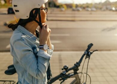A woman wearing a helmet and denim jacket secures her chin strap while standing next to her bicycle on a paved street.