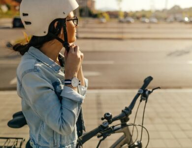 A woman wearing a helmet and denim jacket secures her chin strap while standing next to her bicycle on a paved street.