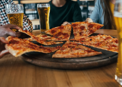 Three people sit at a table with glasses of beer, reaching for slices of pizza from a wooden serving board—a perfect way to enjoy an evening while exploring things to do in Seattle.