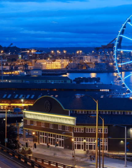 Explore Seattle’s vibrant waterfront at dusk, where a brightly lit Ferris wheel stands out amid city buildings, glowing road light trails, and harbor cranes in the background.