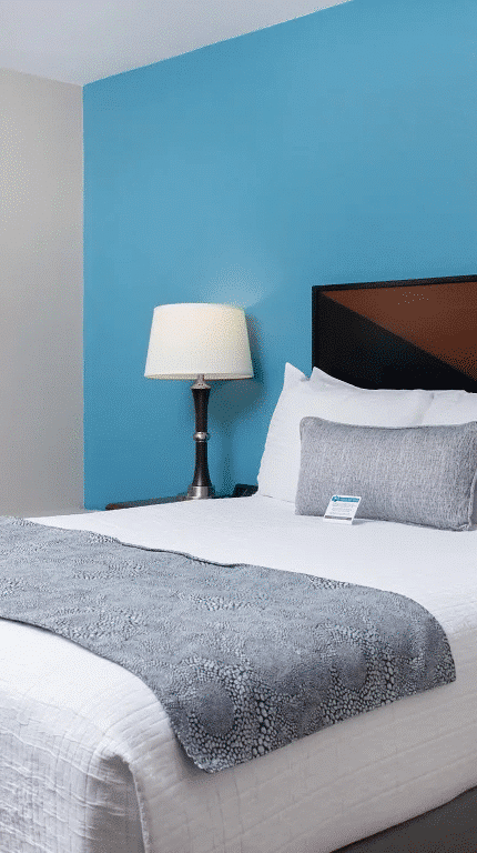 A hotel room with a neatly made bed, blue accent wall, bedside tables with lamps, an armchair by the window, and light-colored curtains.