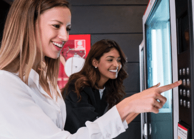 Two women standing in front of a vending machine, one smiling and pressing buttons while the other looks on.