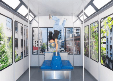 A person does a one-handed handstand on a seat inside a subway car, surrounded by windows showing urban buildings and greenery.