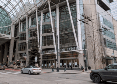 Street view of a large glass and steel convention center with cars at an intersection and pedestrians walking near the entrance.