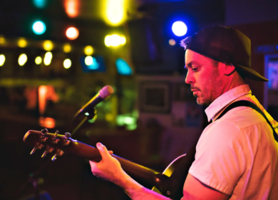 A man wearing a backwards cap plays an acoustic guitar on stage, with colorful lights and a microphone visible in the background.