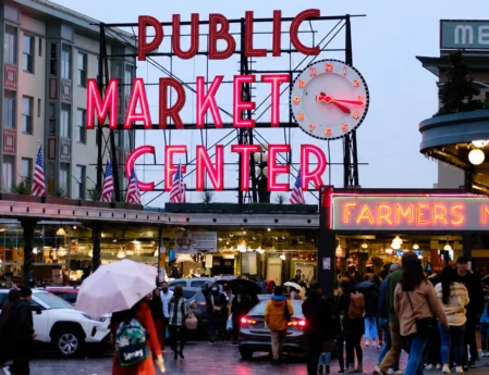 Evening view of iconic neon "Public Market Center" sign and clock above bustling farmers market entrance, cobblestone street and shoppers with umbrellas