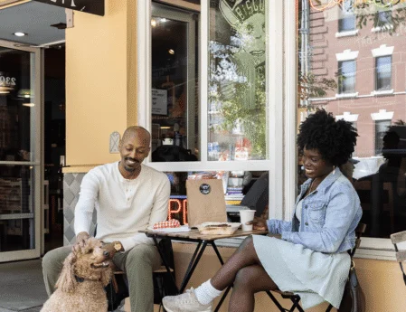 Smiling couple enjoying coffee and pastries at a sunny pavement café table with takeaway bag and friendly dog — breakfast, outdoor seating, pet-friendly hotel nearby