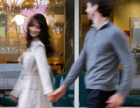 Smiling couple holding hands passing hotel lobby window with cosy seating, pendant lights and contemporary decor visible