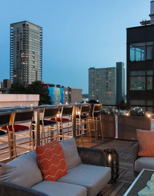Rooftop hotel lounge at dusk with cosy sofas, bar stools, city skyline views, mood lighting and evening drinks and cushions
