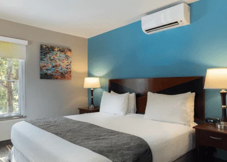 Bright double king bed with crisp white linens, bedside lamps, air-conditioning unit, window with tree view and modern artwork.