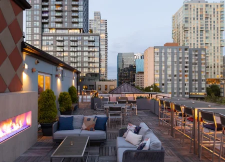 Rooftop terrace with cosy sofas, outdoor fireplace, bar seating and city skyline views at dusk — ideal for drinks and relaxing stays