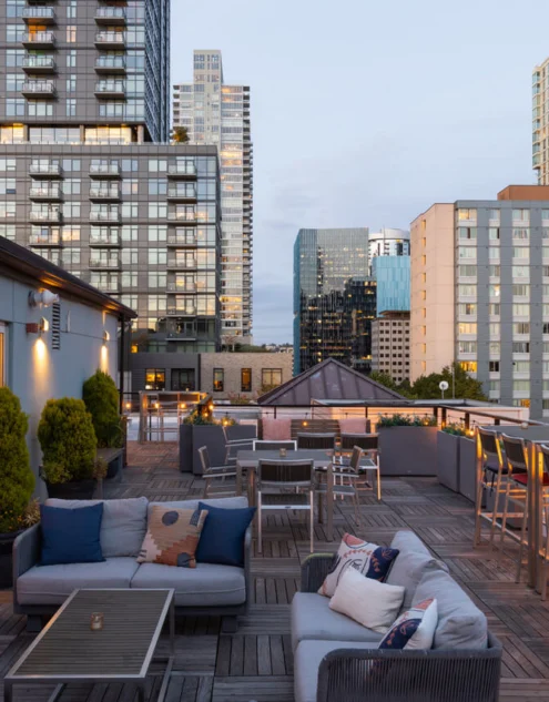 Rooftop terrace with cosy sofas, outdoor fireplace, bar seating and city skyline views at dusk — ideal for drinks and relaxing stays