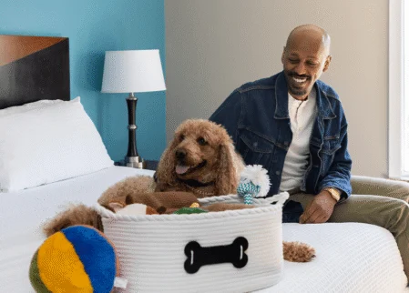 Guest with small brown doodle dog relaxing on a crisp white king bed beside bedside lamp and pet toy basket — pet‑friendly hotel room.