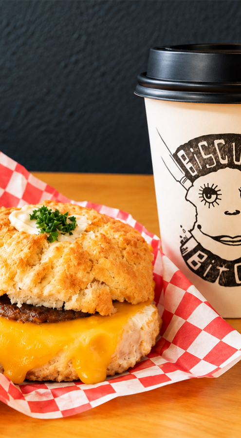 Warm cheddar-topped sausage biscuit on red-check paper beside branded takeaway coffee cup, cosy breakfast at hotel café
