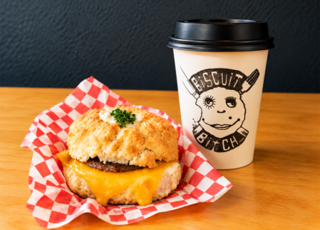 Cheesy sausage breakfast biscuit on red-check paper beside takeaway coffee cup on timber table, inviting hotel breakfast amenity